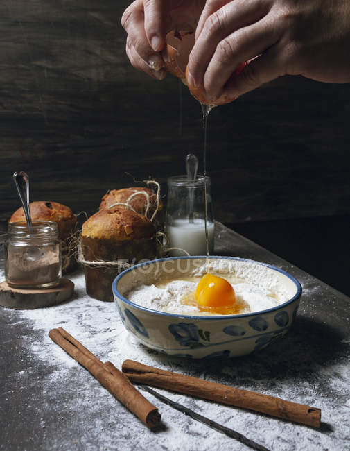Mains de la personne cassant l'œuf dans un bol placé sur une table en bois avec de la cannelle et de la vanille et des gâteaux cuits au four — Photo de stock