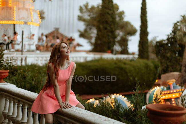 Mujer joven en vestido rosa sentado en la valla en el parque de atracciones - foto de stock