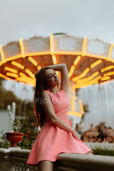 Jovem mulher em vestido rosa sentado na cerca no parque de diversões — Fotografia de Stock
