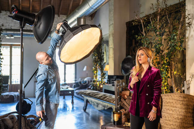 Photographe professionnel réglant des lumières avec femme sensuelle confiante à l'intérieur de la maison de campagne confortable — Photo de stock