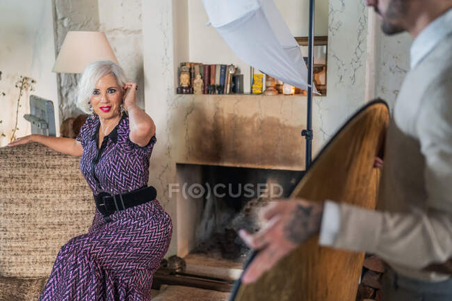 Pensive grauhaarige Schauspielerin in elegantem Sitz neben Softbox und Blick in die Kamera während der Arbeitspause vor verschwommenem Interieur des gemütlichen zeitgenössischen Studios — Stockfoto
