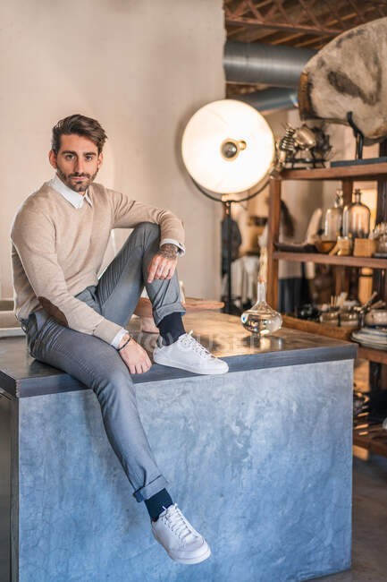 Shooting of stylish adult guy in casual clothes looking away and contemplating while sitting with leg bent on wooden table against small window in light photo studio — Stock Photo