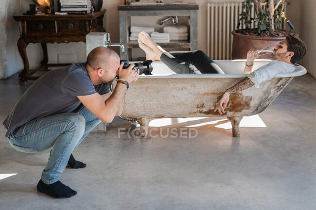 Diligente fotógrafo tomando fotos con cámara de hombre acostado en el baño contra el interior en estilo retro - foto de stock