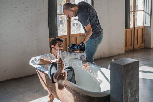 Diligent photographer taking shot with camera of male lying in bath against interior in retro style — Stock Photo