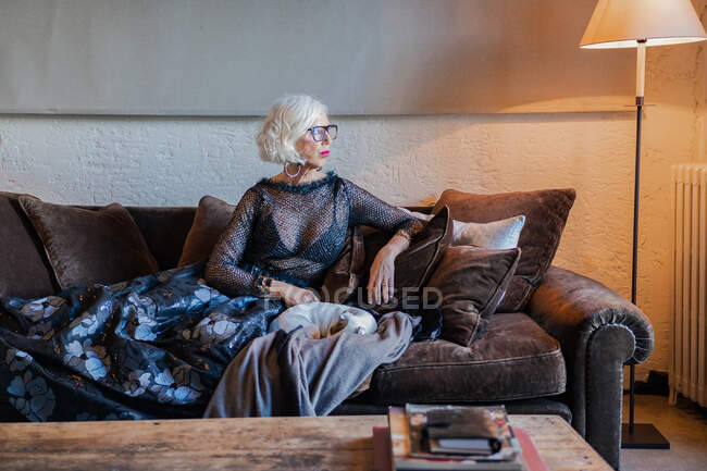 Спокойная уверенная леди в элегантном черном платье смотрит вдаль и мечтает, сидя в мягком коричневом кресле против стареющего деревенского интерьера — стоковое фото