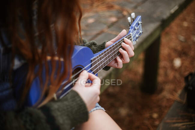 Сверху неузнаваемая брюнетка в повседневной одежде, играющая на маленькой голубой гитаре, сидя одна на деревянной скамейке в осеннем городском парке — стоковое фото