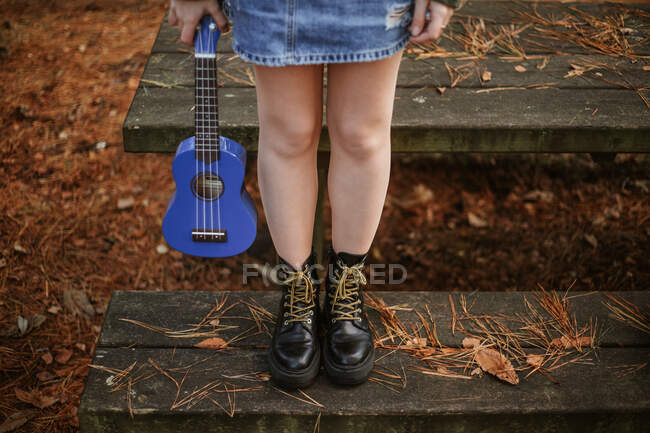 High angle of crop teenager in blue denim skirt and black leather boots standing alone on old wooden bench beside table among brown dry leaves and pine needles and holding small blue guitar in autumn forest — Stock Photo