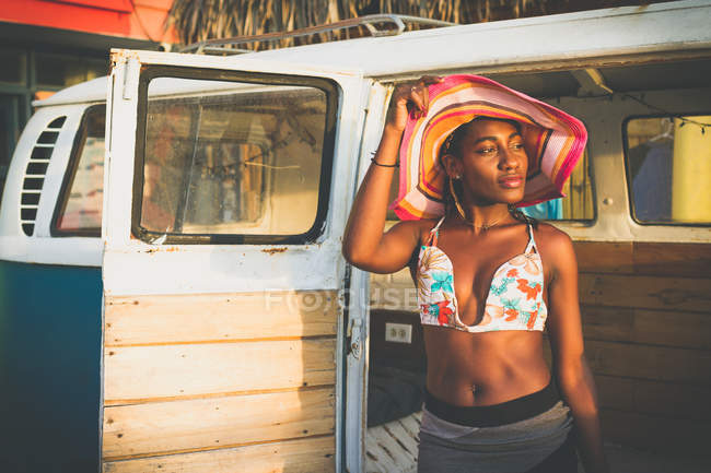 Joven mujer afroamericana confiada en bikini colorido superior tocando sombrero de rayas de ala ancha y mirando hacia otro lado - foto de stock