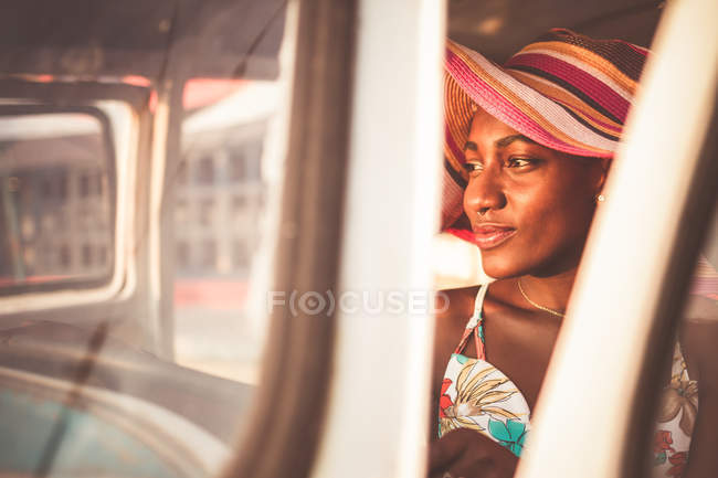 Positiva jovem afro-americana em chapéu listrado colorido sentado ao volante do carro velho e olhando para longe — Fotografia de Stock