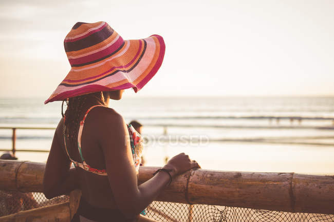 Side view of relaxed young African American woman in colorful wide brimmed striped hat and bikini top standing at wooden fence on beach — Stock Photo