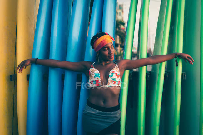 Young African American woman wearing colorful bikini top and headband spreading arms while standing back to colorful surf boards — Stock Photo