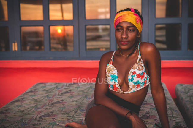 Pensive calm African American woman wearing colorful bikini top and headband sitting on lounge in room with red floor and red sunset through window in background — Stock Photo