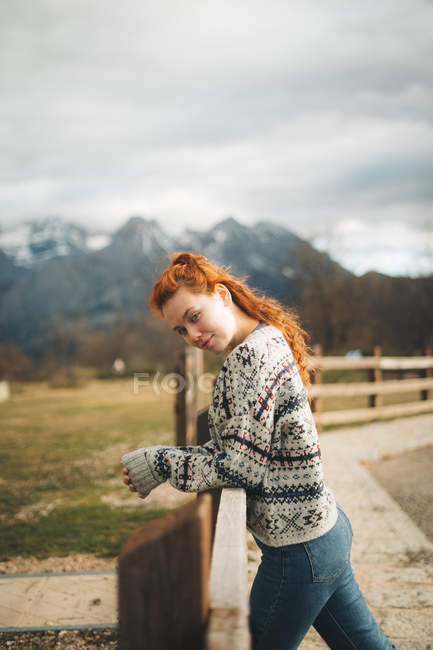 Vista lateral de mujer encantadora con confianza con el pelo rojo en suéter caliente apoyado en valla de madera a lo largo de camino en la montaña - foto de stock
