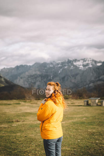 Vista posteriore di donna sorridente in giacca gialla calda guardando la fotocamera sopra la spalla in prato verde erba circondato da montagne innevate — Foto stock