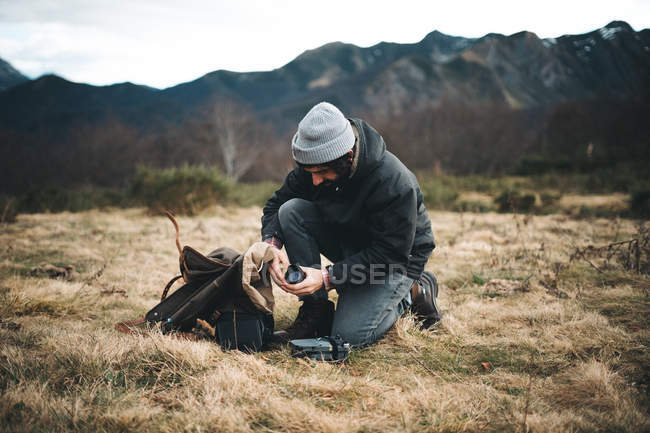 Vue latérale de l'homme en chapeau et veste chaude prenant accessoires de la caméra dans la prairie avec herbe sèche — Photo de stock