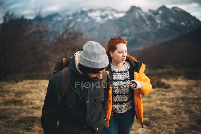 Amorous homme avec sac à dos tenant la main avec belle femme rousse dans classé avec de l'herbe sèche près des montagnes par temps nuageux — Photo de stock