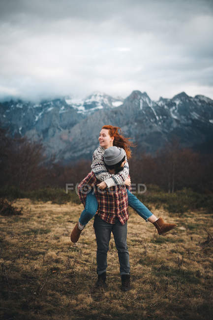 Preciosa pareja abrazando y pasando tiempo juntos a cuestas y viajando en el césped con hierba seca cerca de las montañas en el día nublado - foto de stock