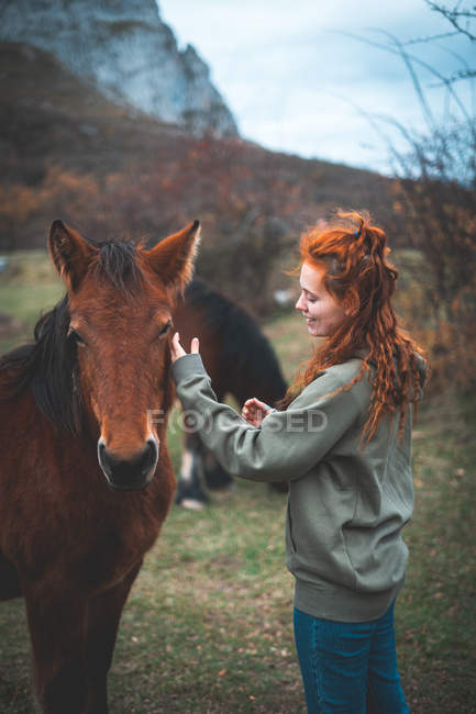 Вид сбоку улыбающейся женщины с длинными рыжими волосами в капюшоне, гладящей коричневую лошадь с черным маном в горном пасте — стоковое фото