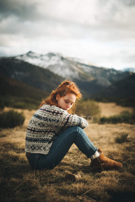 Vista laterale di donna affascinante pensosa con i capelli rossi in maglione caldo seduto con gli occhi chiusi sul prato in montagna in giornata grigia — Foto stock
