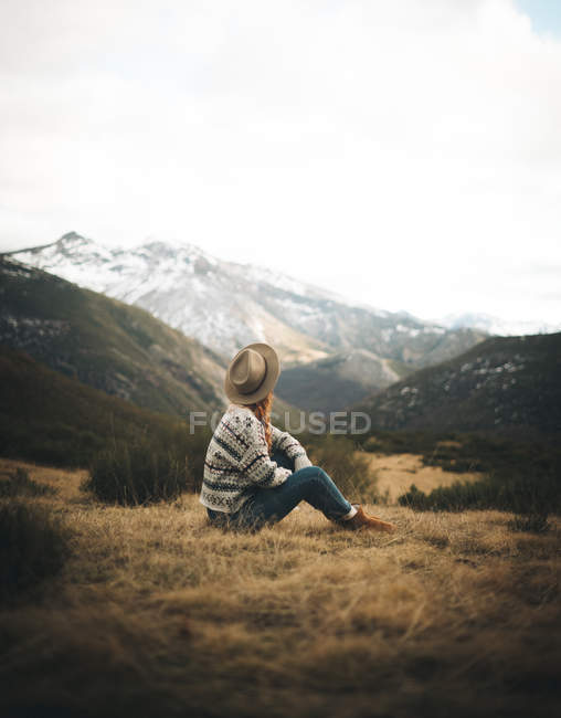 Vue latérale d'une femme de charme méconnaissable et coûteuse aux cheveux roux dans un pull chaud assis sur la pelouse en montagne par temps gris — Photo de stock