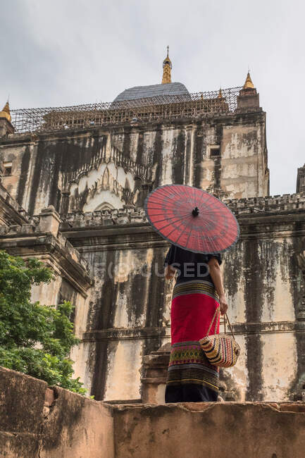 Female traveler in traditional clothes with red umbrella standing next to old building — Stock Photo