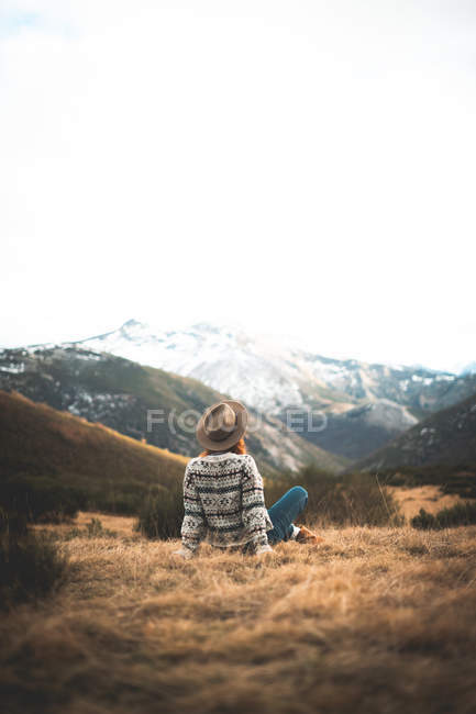 Vista posterior de mujer encantadora pensativa irreconocible con el pelo rojo en suéter caliente sentado en el césped en la montaña en día gris - foto de stock