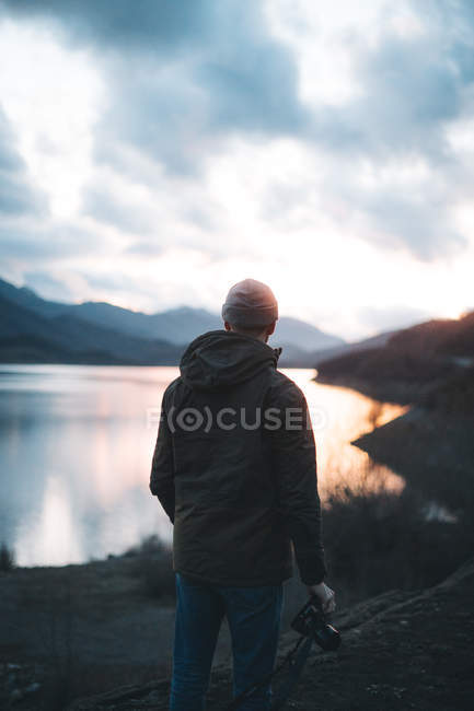 Back view of traveler in jacket in hat and shirt with camera looking back on stony mountains with tree — Stock Photo
