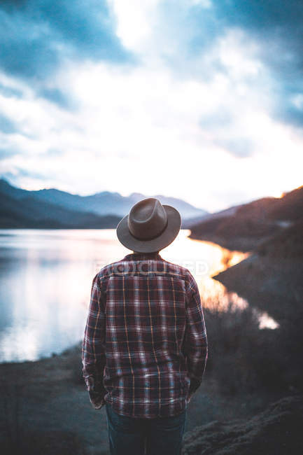 Visão traseira do viajante de jaqueta em chapéu e camisa em montanhas pedregosas durante o pôr do sol — Fotografia de Stock