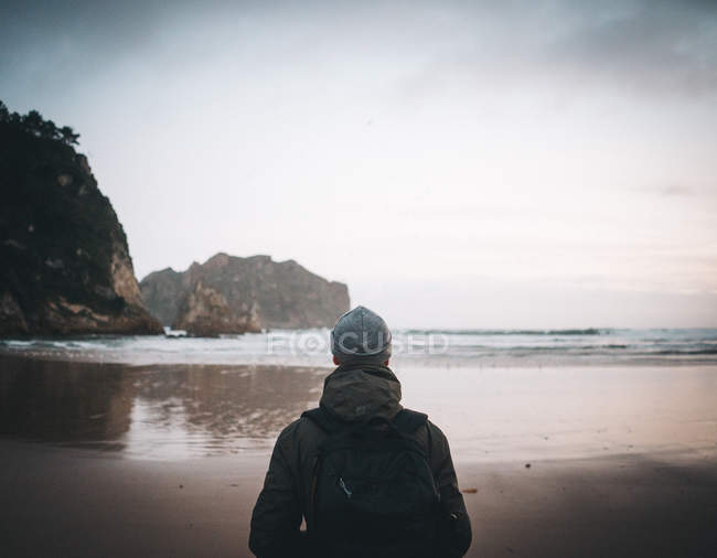 Vista trasera de viajero anónimo en chaqueta con mochila contemplando vistas de la costa rodeada de montaña - foto de stock