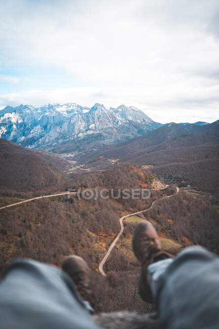 Crop traveler in brown boots sitting with legs dangling on edge on rock and enjoying breathtaking view of forest and road — Stock Photo
