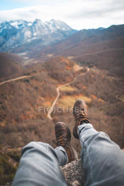 Viajero de cultivos en botas marrones sentado con las piernas colgando en el borde de la roca y disfrutar de impresionantes vistas del bosque y la carretera - foto de stock