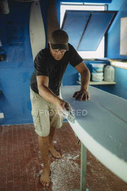 Facharbeiter glättet barfuß weißes Surfbrett in Werkstatt mit blauen Wänden — Stockfoto