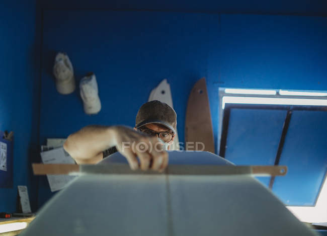 Концентрированный квалифицированный рабочий в защитной маске, выравнивающий белую доску для серфинга в мастерской с голубыми стенами — стоковое фото