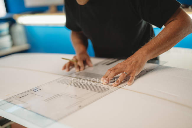 Zugeschnittenes Bild eines professionellen Männchens mit Bleistift in der Hand mit Messwerkzeug beim Herstellen eines Surfbretts in der Werkstatt — Stockfoto