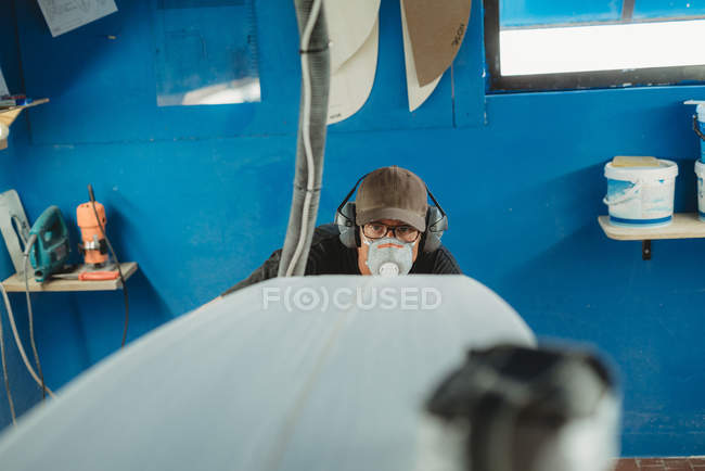 Handwerker mit Schutzmaske und Kopfhörer bastelt Surfbrett in kleiner Werkstatt mit blauen Wänden — Stockfoto