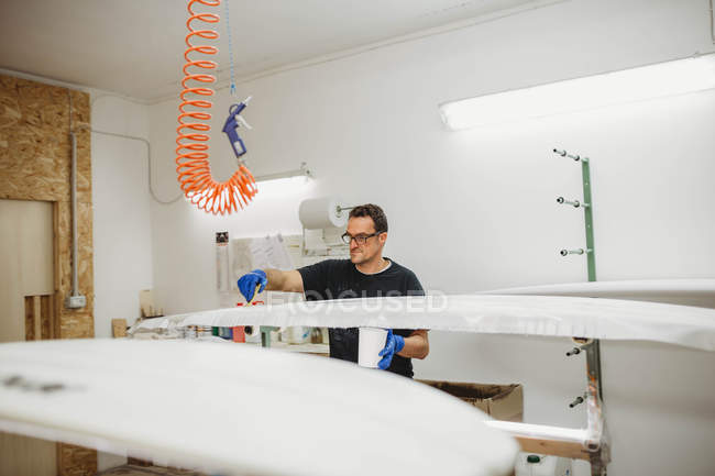 Professional adult man in protective gloves painting surf board in white color while working in workshop — Stock Photo