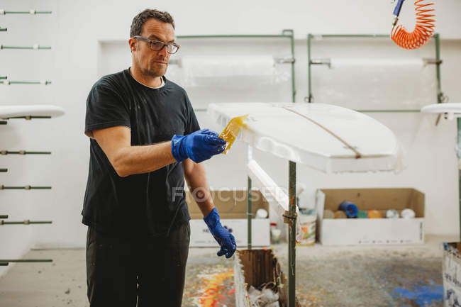 Ouvrier adulte en gants et lunettes de protection peignant la planche de surf blanche tout en travaillant dans un petit atelier — Photo de stock