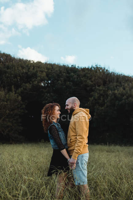 Hombre con capucha amarilla tomados de la mano a una novia feliz en vestido y chaleco de mezclilla mientras están de pie juntos en el prado - foto de stock