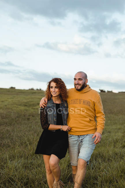 Cheerful romantic young couple holding hands and walking on green field at seaside at sunset time with cloudy sky — Stock Photo