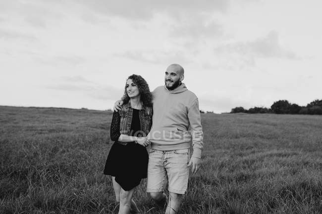 Alegre casal jovem romântico de mãos dadas e caminhando no campo verde à beira-mar ao pôr-do-sol com céu nublado — Fotografia de Stock