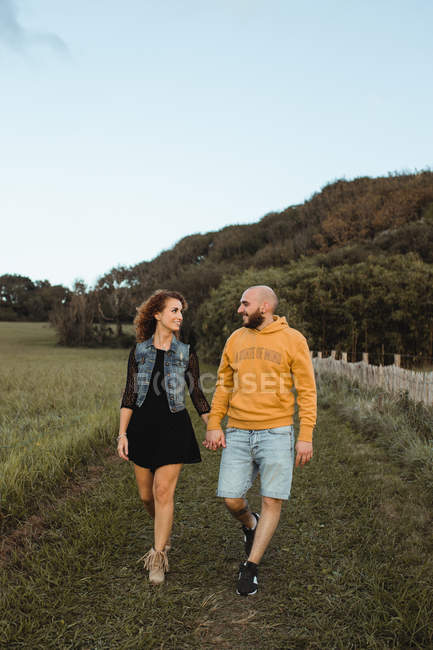 Весела романтична молода пара тримає руки і ходить на зеленому полі на узбережжі під час заходу сонця з хмарним небом — стокове фото