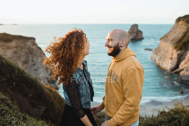 Вид сбоку счастливой пары, держащейся за руки и разговаривающей, стоя на скалистом берегу моря и проводя солнечный вечер вместе — стоковое фото