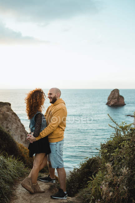Side view of happy couple holding hands and talking while standing on rocky seashore and spending sunny evening together — Stock Photo