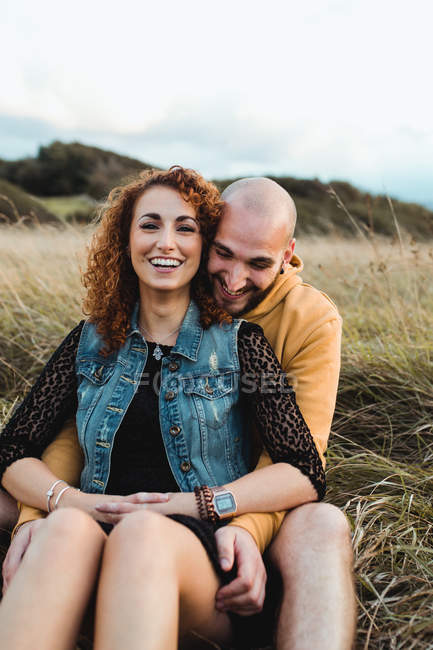 Jeune homme en sweat à capuche jaune embrassant petite amie en robe et gilet en denim tout en se tenant près de la clôture ensemble sur la prairie — Photo de stock