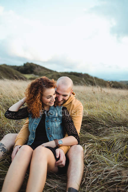 Jeune homme en sweat à capuche jaune embrassant petite amie en robe et gilet en denim tout en se tenant près de la clôture ensemble sur la prairie — Photo de stock