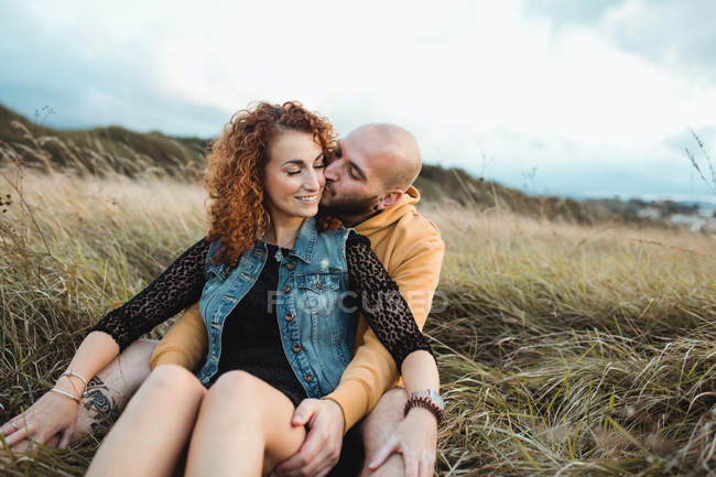 Joven con capucha amarilla abrazando a su novia en vestido y chaleco de mezclilla mientras está de pie cerca de la valla juntos en el prado - foto de stock