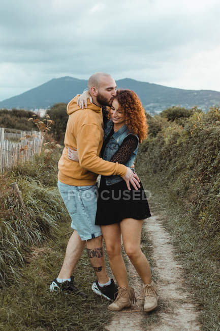 Amoroso Jovem de capuz amarelo abraçando a namorada em vestido e colete de ganga enquanto estava perto de cerca juntos no prado — Fotografia de Stock