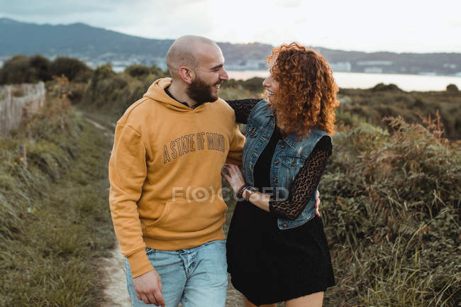 Loving couple walking together on walkway along green farm at seaside with mountain and city buildings in background — Stock Photo