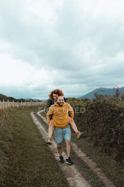 Счастливый хипстер катает подружку на спине во время прогулки по сельской дороге между зеленой фермой и морским побережьем в пасмурный день — стоковое фото
