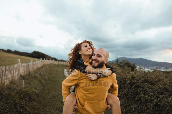 Happy hipster man giving girlfriend piggyback ride while walking on rural road between green farm and sea coast in cloudy day — Stock Photo